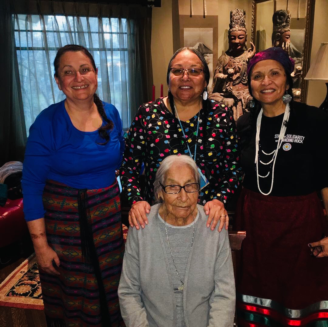 Four women, three standing and one seated, are smiling in a cozy room. The seated woman, with glasses, conveys warmth. Decor includes statues, evoking a cultural ambiance.