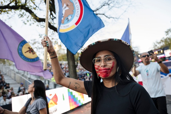 A woman wearing a large hat and glasses, holding a flag while displaying red paint on her face. In the background, other individuals are participating in an event with colorful flags and banners visible.
